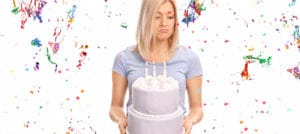 Sad young woman holding a birthday cake and looking down with a bunch of confetti streamers flying around her isolated on white background